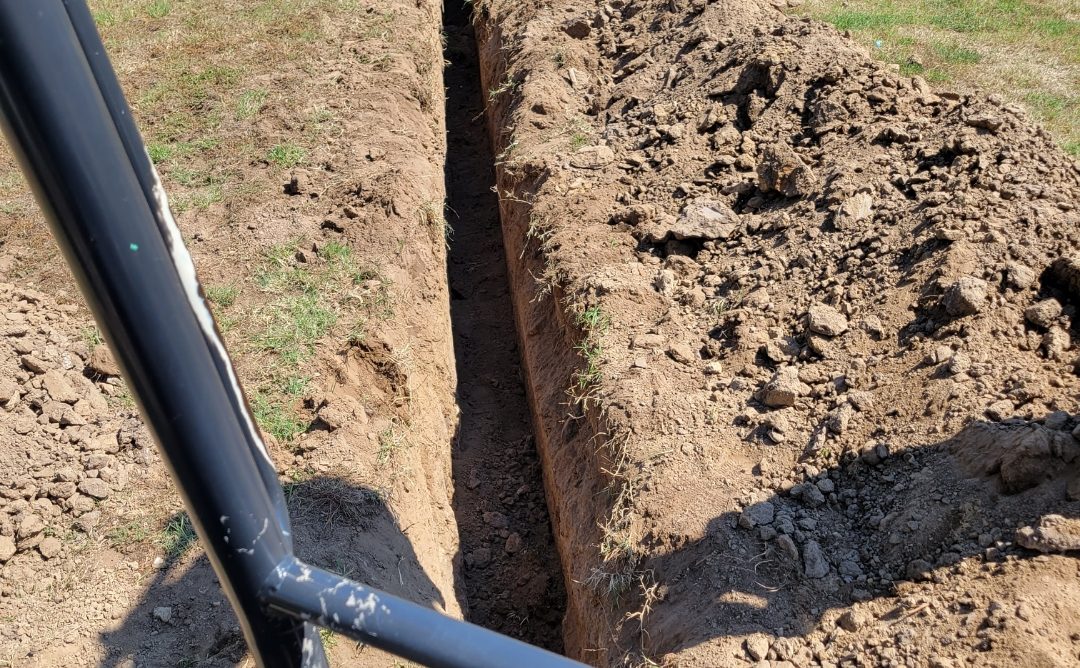 A narrow trench dug in the ground beside a black pipe.